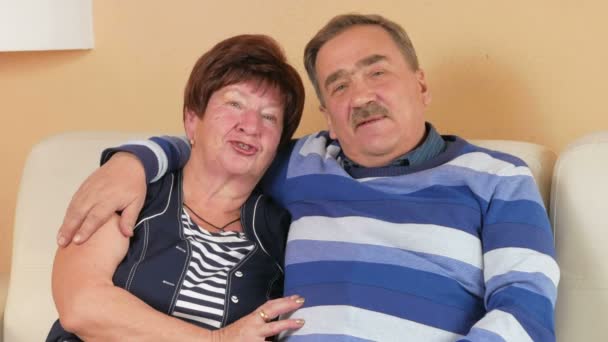 Happy senior man embracing his beloved wife on a sofa in a beautiful house. They talk and laugh. Looking at the camera with a smile — Stock Video