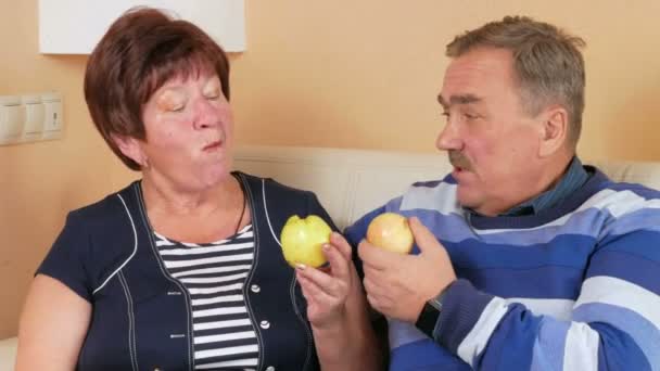 Casal sênior descansando em casa no sofá e comer maçãs. Na mesa está um prato de frutas diferentes. O marido e a esposa falam um com o outro . — Vídeo de Stock