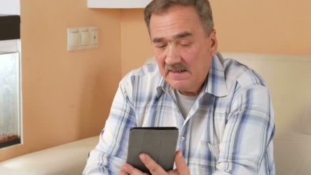 Man senior mustasch läsa nyheter på tablet PC hemma. Han sitter i soffan nära akvariet och dricka kaffe. — Stockvideo