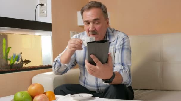 Man senior mustasch läsa nyheter på tablet PC hemma. Han sitter i soffan nära akvariet och dricka kaffe. — Stockvideo