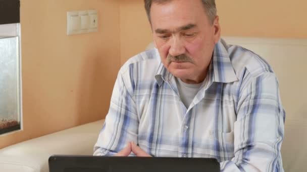 Старший чоловік вуса читає новини на ноутбуці вдома. Він сидить на дивані біля акваріума і дивиться на екран комп'ютера . — стокове відео