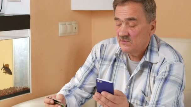 Старший мужчина с усами делает покупку с кредитной карты интернет-магазина. Он сидел дома на диване и вводит информацию о карточке, чтобы позвонить — стоковое видео