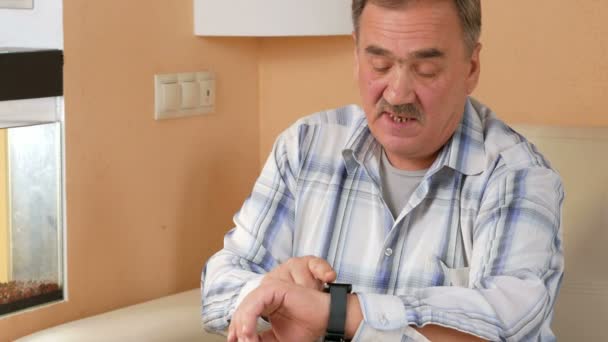 Senior man snor controleert berichten op de smart watch thuis. Hij zit op de Bank in de buurt van het aquarium en het bericht spreekt. — Stockvideo