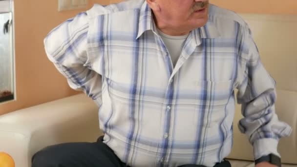 Старший чоловік з вусами не міг вийти з дивана через біль у спині. Він тримається за талію руками і страждає болем вдома — стокове відео