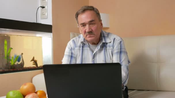 Старший чоловік вуса читає новини на ноутбуці вдома. Він сидить на дивані біля акваріума і дивиться на екран комп'ютера . — стокове відео
