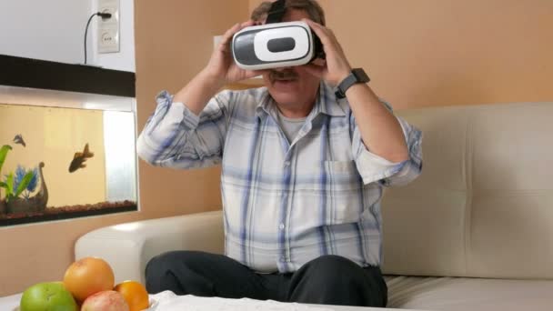 Hombre mayor con bigote está sentado con un casco de realidad virtual en el sofá en casa. Él se pregunta lo que ven y tratar de tocar sus manos a los elementos virtuales — Vídeos de Stock