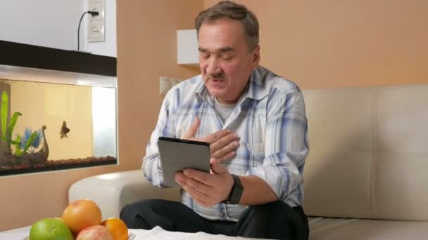 Uomo anziano con i baffi in tosse e comunicare con un medico in una videoconferenza. Medico su un tablet on-line fa raccomandazioni al paziente a casa — Video Stock