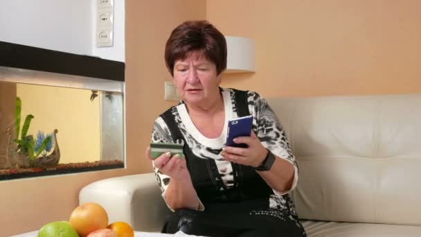 Kredi kartı ve telefon ile çevrimiçi alışveriş yapmak kadın yaşlı. Dikkatli bir şekilde telefonunuza harita verileri tanıtır. Akvaryum ayakta evde kanepede oturan — Stok video
