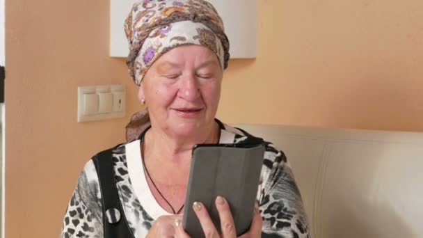 Senior Kvinna pratar på tablet Pc med en flickvän. Hon sitter i soffan hemma nära akvariet. Han ler och berättar om livet. — Stockvideo
