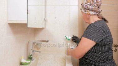 Üst düzey kadın otel odasında banyo lavabo temizler. O eldivenleri ve temizleyici kullanır. Bir sünger yıkama elinde