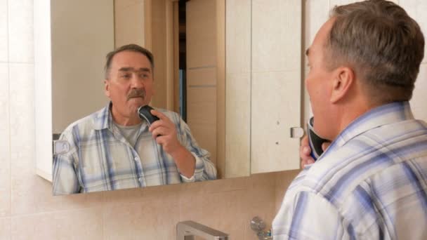 Homem envelhecido barbear navalha elétrica no banheiro. Ele olha no espelho e cuidadosamente rapa todo o cabelo dela. Deixa apenas um bigode. . — Vídeo de Stock
