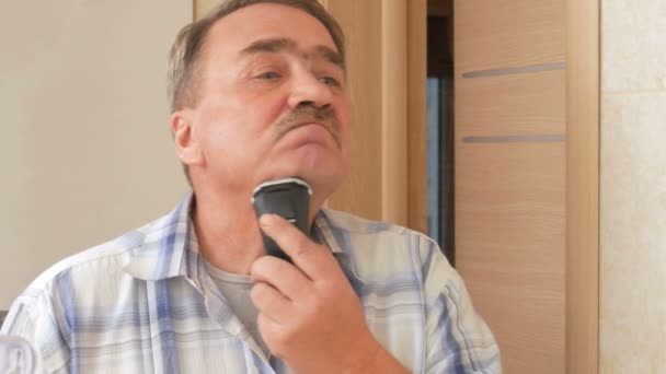 Homme vieux rasage rasoir électrique dans la salle de bain. Il regarde dans le miroir et se rase soigneusement tous ses cheveux. Il ne laisse qu'une moustache . — Video