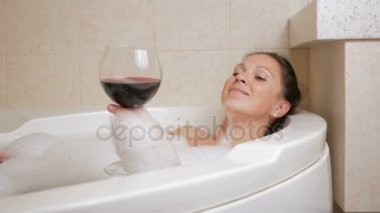 Bir kadeh şarap ile köpük banyosu alarak güzel kız. Bir büyük beyaz banyo ve yüzünde sevinç. gevşeme kavramı