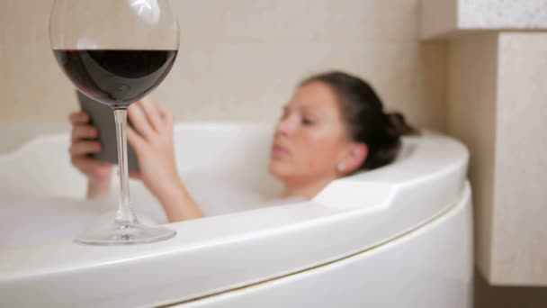 Belle fille prenant un bain avec mousse et tablette PC. Un grand bain blanc et un verre de vin rouge. Cliquer sur l'assiette et boire du vin — Video