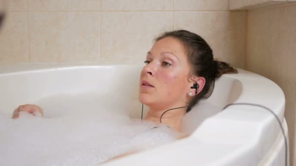 Όμορφη κοπέλα Λαμβάνοντας ένα μπάνιο με αφρό και να ακούτε μουσική με ακουστικά. Ένα μεγάλο λευκό μπάνιο και ένα ποτήρι κόκκινο κρασί. Τραγουδήστε μαζί με ένα αγαπημένο τραγούδι. έννοια της χαλάρωσης — Αρχείο Βίντεο