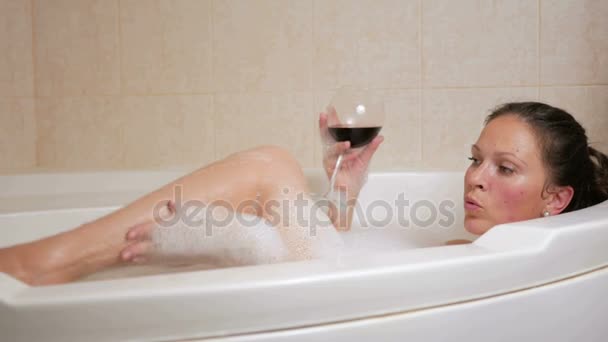Красивая девушка принимает ванну с пеной и бокалом вина. Большая белая ванна и радость на его лице. релаксация — стоковое видео