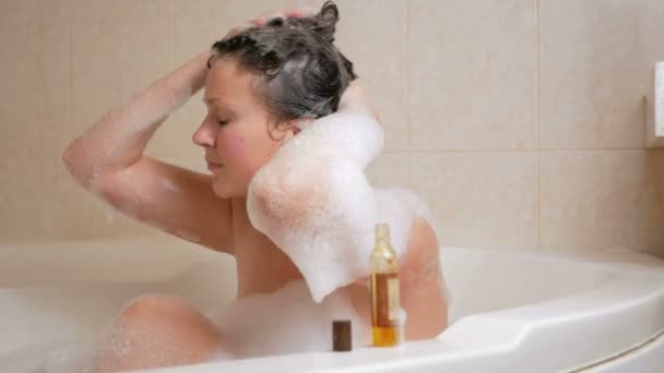 A young girl takes a bath with foam and washing hair with shampoo. A large white bath and a small bottle of shampoo at the hotel. — Stock Video