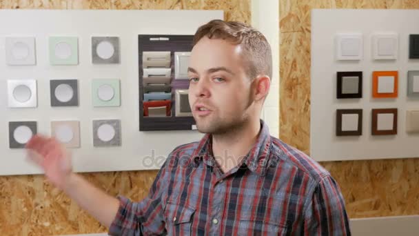 Un jeune homme raconte et montre des interrupteurs de lumière de différentes couleurs et formes. Les échantillons sont présentés au stand. Magasiner électriciens — Video