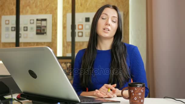 Aantrekkelijke vrouw die werkt in een kantoor op de computer. Ze ziet er in de camera in gesprek met een klant. Lang donker haar en blauwe jurk — Stockvideo