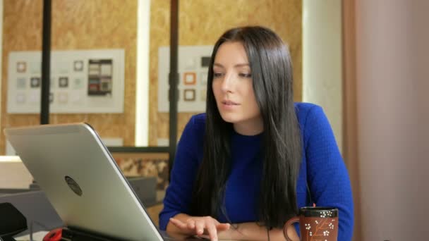 Привлекательная женщина, работающая в офисе за компьютером. Она смотрит в камеру и улыбается. Длинные темные волосы и синее платье — стоковое видео
