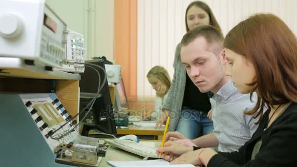 Un gruppo di studenti che studiano all'università come ingegnere. Siediti intorno allo stand con l'attrezzatura. Diverse persone prendono consiglio tra di loro e dimostrano il loro punto di vista. Registrare i dati . — Video Stock