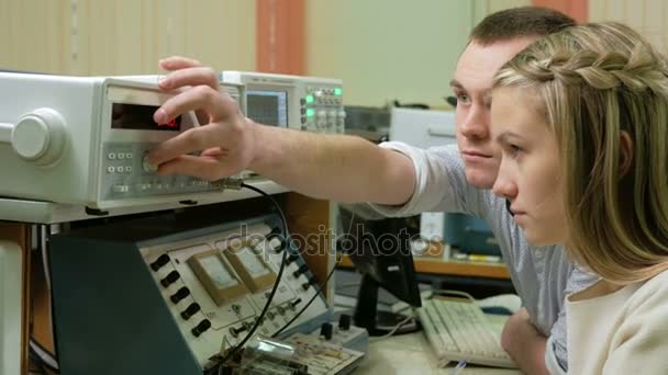 Una joven estudiante de ingeniería que trabaja con un hombre en el laboratorio con un equipo de medición eléctrico. Giran cuidadosamente el reloj y los datos registrados — Vídeo de stock