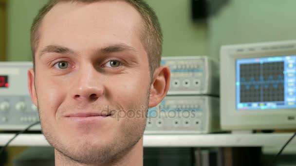 Молодой привлекательный мужчина в комнате с электрическим оборудованием улыбается в камеру. На фоне графика на осциллографе. Концепция счастливого ученого — стоковое видео
