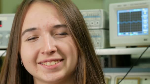 Mujer atractiva joven en la habitación con equipo eléctrico sonriendo a la cámara. Sobre el fondo de un gráfico en el osciloscopio. El concepto de un científico feliz — Vídeos de Stock