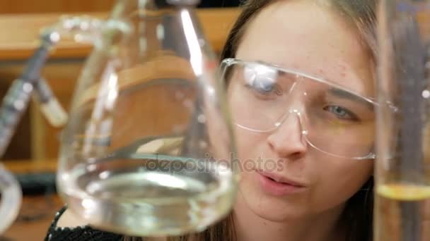 Молодая студентка работает с химикатами и пишет результаты в университете. Очки и халат. Стаканы и колбы с жидкостью . — стоковое видео