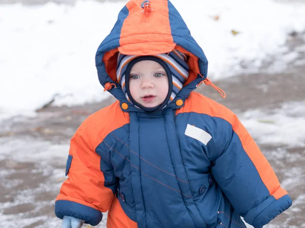 Привабливий хлопчик грає з першим снігом. Він посміхається і виглядає сніговиком. Товстий синьо-помаранчевий комбінезон яскраво смугастий капелюх на річній дитині . Стокове Фото