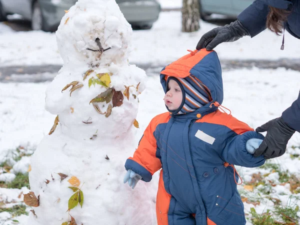 Attraktiver kleiner Junge beim Spielen mit dem ersten Schnee. Er lächelt und sieht aus wie ein Schneemann. dicker blau-orangefarbener Overall mit hellen Streifen auf einem einjährigen Kind. lizenzfreie Stockbilder