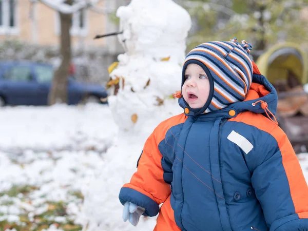 Menino atraente brincando com a primeira neve. Ele sorri e parece boneco de neve. Macacão azul-laranja grosso chapéu listrado brilhante em uma criança de ano . Fotografia De Stock