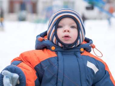 İlk kar ile oynayan erkek çekici bebek. O gülümser ve kardan adam görünüyor. Bir yaşındaki çocuk kalın mavi-turuncu tulum parlak çizgili şapkası.