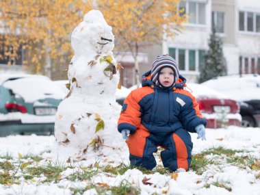 İlk kar ile oynayan erkek çekici bebek. O gülümser ve kardan adam görünüyor. Bir yaşındaki çocuk kalın mavi-turuncu tulum parlak çizgili şapkası.