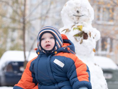 İlk kar ile oynayan erkek çekici bebek. O gülümser ve kardan adam görünüyor. Bir yaşındaki çocuk kalın mavi-turuncu tulum parlak çizgili şapkası.