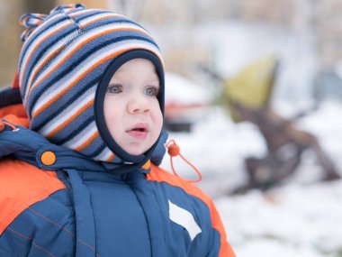 İlk kar ile oynayan erkek çekici bebek. O gülümser ve kardan adam görünüyor. Bir yaşındaki çocuk kalın mavi-turuncu tulum parlak çizgili şapkası.