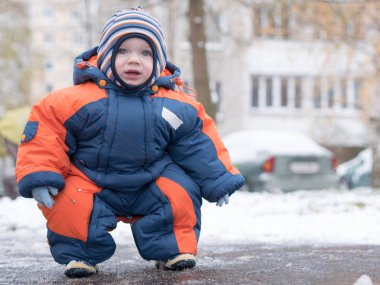 İlk kar ile oynayan erkek çekici bebek. O gülümser ve kardan adam görünüyor. Bir yaşındaki çocuk kalın mavi-turuncu tulum parlak çizgili şapkası.