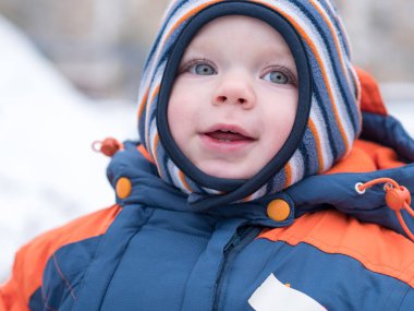İlk kar ile oynayan erkek çekici bebek. O gülümser ve kardan adam görünüyor. Bir yaşındaki çocuk kalın mavi-turuncu tulum parlak çizgili şapkası.