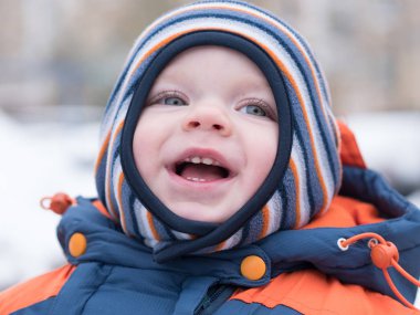 İlk kar ile oynayan erkek çekici bebek. O gülümser ve kardan adam görünüyor. Bir yaşındaki çocuk kalın mavi-turuncu tulum parlak çizgili şapkası.
