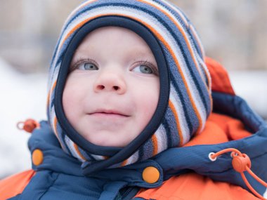 İlk kar ile oynayan erkek çekici bebek. O gülümser ve kardan adam görünüyor. Bir yaşındaki çocuk kalın mavi-turuncu tulum parlak çizgili şapkası.