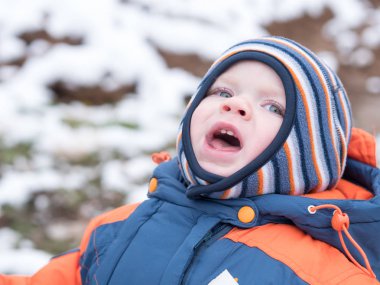 İlk kar ile oynayan erkek çekici bebek. O gülümser ve kardan adam görünüyor. Bir yaşındaki çocuk kalın mavi-turuncu tulum parlak çizgili şapkası.