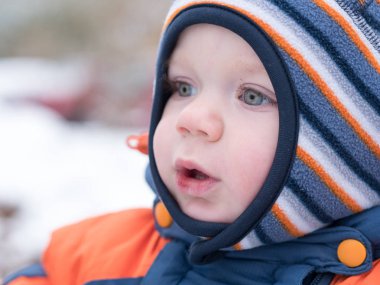 İlk kar ile oynayan erkek çekici bebek. O gülümser ve kardan adam görünüyor. Bir yaşındaki çocuk kalın mavi-turuncu tulum parlak çizgili şapkası.