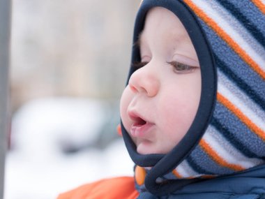İlk kar ile oynayan erkek çekici bebek. O gülümser ve kardan adam görünüyor. Bir yaşındaki çocuk kalın mavi-turuncu tulum parlak çizgili şapkası.