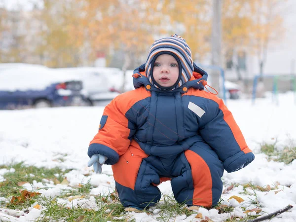 Menino atraente brincando com a primeira neve. Ele sorri e parece boneco de neve. Macacão azul-laranja grosso chapéu listrado brilhante em uma criança de ano . — Fotografia de Stock