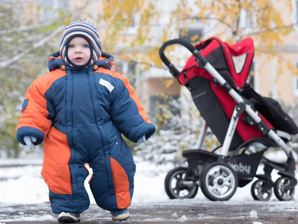 Attraktiva pojke leker med den första snön. Han ler och ser snögubbe. Tjock blå-orange overall ljusa randiga hatten på ett år gammalt barn. — Stockfoto