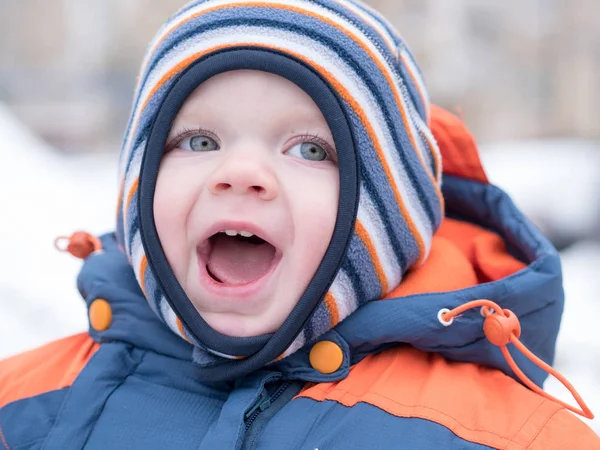 İlk kar ile oynayan erkek çekici bebek. O gülümser ve kardan adam görünüyor. Bir yaşındaki çocuk kalın mavi-turuncu tulum parlak çizgili şapkası.