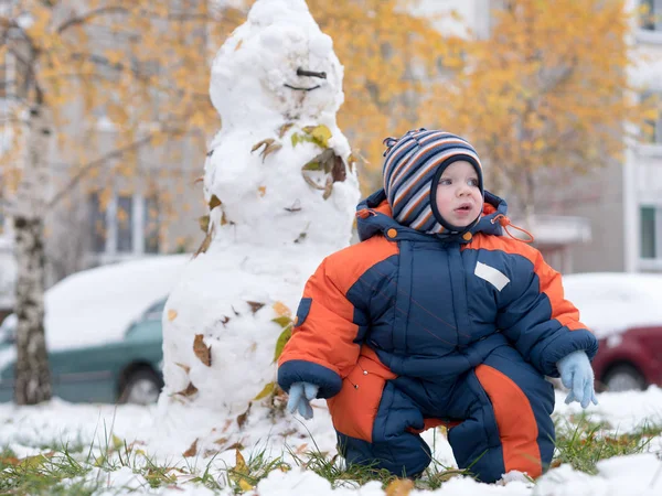 Attraktiver kleiner Junge beim Spielen mit dem ersten Schnee. Er lächelt und sieht aus wie ein Schneemann. dicker blau-orangefarbener Overall mit hellen Streifen auf einem einjährigen Kind. Stockbild