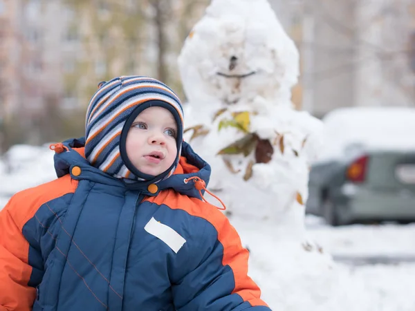 Menino atraente brincando com a primeira neve. Ele sorri e parece boneco de neve. Macacão azul-laranja grosso chapéu listrado brilhante em uma criança de ano . Imagens Royalty-Free