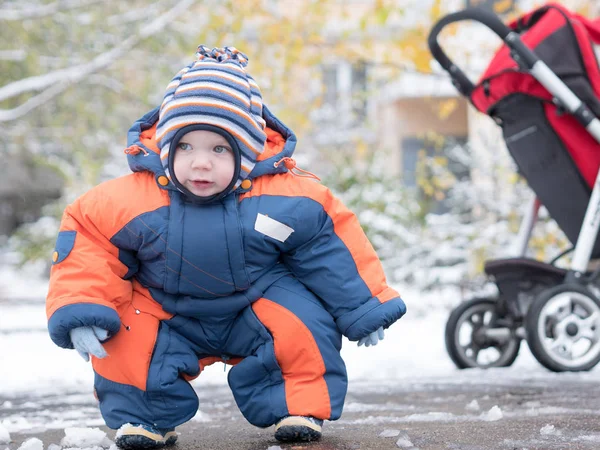最初の雪で遊んで魅力的な男の子。彼の笑顔し、雪だるまに見えます。歳子にジャンプ スーツ ブルー オレンジ厚さ明るいストライプ帽子. ストック画像
