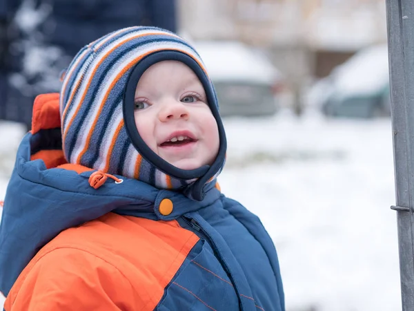 Menino atraente brincando com a primeira neve. Ele sorri e parece boneco de neve. Macacão azul-laranja grosso chapéu listrado brilhante em uma criança de ano . Fotografias De Stock Royalty-Free